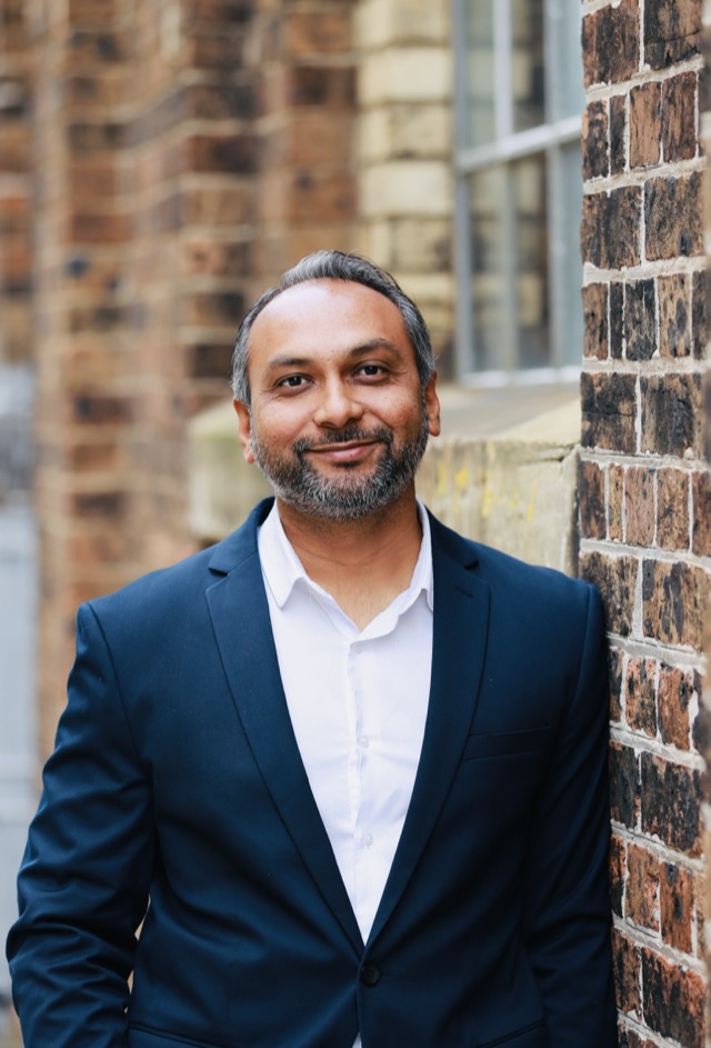 Anit Patel, business coach, smiling in a navy blazer against a brick wall